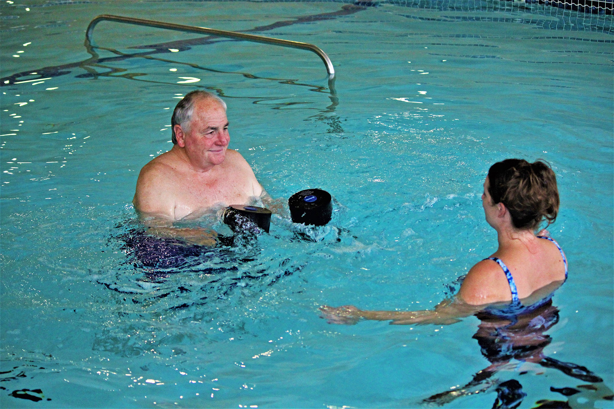 Older man performing aquatic resistance exercises with a therapist in a swimming pool.